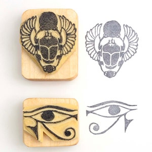 May include: Two wooden stamps with black ink designs. One stamp features a scarab beetle with wings, and the other depicts the Eye of Horus. The stamped impressions are next to the stamps.