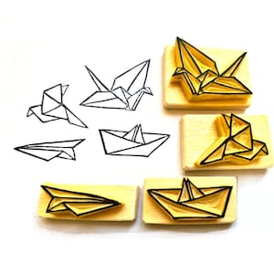 May include: Origami-themed stamps and line drawings. The stamps feature paper crane, paper boat, and paper airplane designs. The line drawings show the same origami figures. The stamps are mounted on rectangular wooden blocks, with black outlines and yellow interiors.