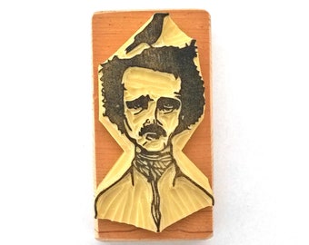Puede incluir: Un sello de madera con una imagen en blanco y negro de Edgar Allan Poe. La imagen es de la cabeza y los hombros de Poe, con su cabello y barba visibles. El sello está sobre una base de madera marrón claro.
