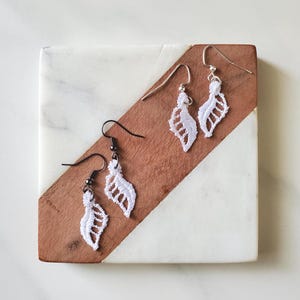 May include: Three pairs of white lace leaf earrings with different hook colors: silver and black. The earrings are displayed on a square marble and wood coaster. The background is a white marble surface.