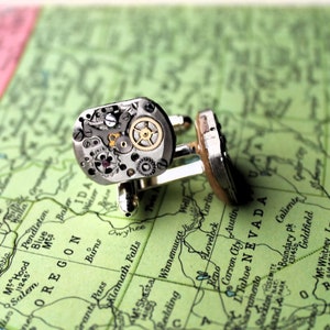 May include: A pair of silver cufflinks featuring intricate watch movement components. The cufflinks showcase gears, jewels, and other mechanical parts, set against a map background. The cufflinks are a unique accessory.