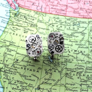 Peut inclure: Deux boutons de manchette rectangulaires argentés ornés d'engrenages de montre complexes et de petits bijoux rouges. Ils sont présentés sur une carte vintage des États-Unis, pour un style steampunk.