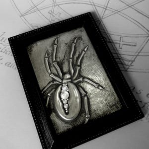 May include: A framed, silver-toned spider sculpture with a detailed, textured body and legs. The spider's abdomen features a row of clear, round embellishments. The black frame has a dotted border, and the background is a textured, gray surface.