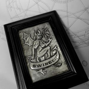 Puede incluir: Escultura en relieve metálico enmarcada que presenta una figura alada y con cuernos con la palabra "SWINGER" en una pancarta. La obra de arte está enmarcada en un marco de madera negro. El fondo tiene una apariencia texturizada.