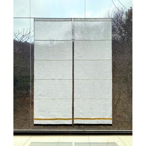 Pode incluir: Duas cortinas romanas brancas com costuras horizontais penduradas em frente a uma janela. As cortinas têm uma fina faixa dourada na parte inferior, em um fundo de árvores e céu.