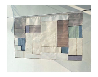 May include: A rectangular wall hanging with a geometric design. The design is made up of various colored squares and rectangles, including beige, brown, blue, green, and purple. The squares and rectangles are arranged in a random pattern.