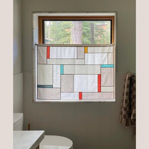 May include: A geometric patterned window curtain with a neutral color palette of gray, white, and beige. The design incorporates rectangular and square shapes with accents of red, teal, and yellow. The curtain is hanging in a bathroom setting.