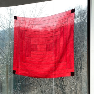May include: A red fabric wall hanging with a geometric pattern of squares and rectangles. The fabric is textured and has a subtle ombre effect.