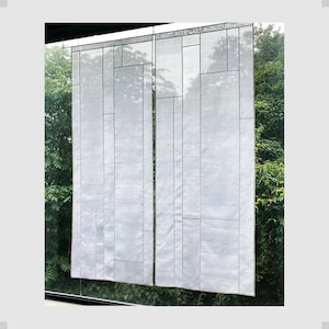 May include: Two white fabric panels with a geometric pattern of squares and rectangles, hanging in a window with green foliage visible behind them.