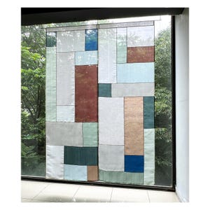 以下が含まれることがあります： パッチワークデザインの長方形の窓の覆い。白、グレー、青、緑、茶色のさまざまな長方形の形が特徴です。緑の木々が見える窓の前に覆いが吊るされています。