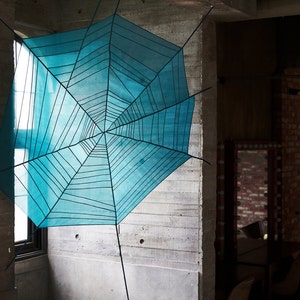May include: A large, turquoise spiderweb art installation suspended in a room with concrete walls. The web is made of thin black lines and is attached to the ceiling and walls. The room has a window and a brick wall.
