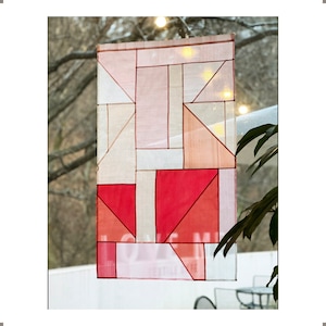 May include: A geometric pattern of pink and red squares and triangles on a white fabric background. The fabric is hanging in a window.