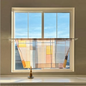 May include: A window with a sheer curtain featuring a geometric design in shades of yellow, orange, brown, and blue. A decorative candle sits on a small plate in front of the window, which has a clear blue sky in the background.