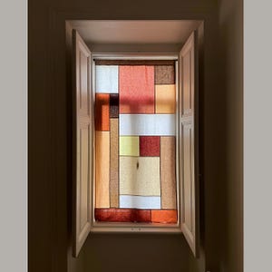 以下が含まれることがあります： 白いシャッターとカラフルな幾何学模様のカーテンが付いた窓。カーテンは、赤、オレンジ、黄色、茶色の長方形と正方形の形を特徴としています。窓はニュートラルな色調の壁に設置されています。