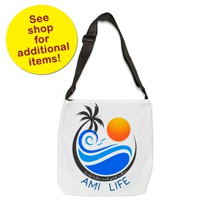 K&ouml;nnte beinhalten: Wei&szlig;e Canvas-Tragetasche mit einem verstellbaren schwarzen Schultergurt. Die Tasche zeigt eine farbenfrohe Grafik mit einer Palme, Sonne und Wellen mit dem Text "Gulf tides and good vibes AMI LIFE".