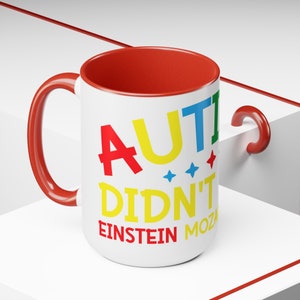 May include: White ceramic coffee mug with a red handle and red rim. The mug has the text "AUTI DIDN'T" in colorful letters, with "EINSTEIN" and "MOZART" in black letters below.