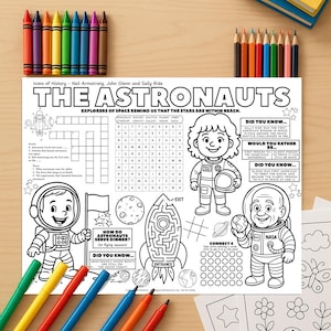 May include: An astronaut-themed activity book with the title "The Astronauts." Features a crossword, maze, and connect-four game. Colouring pencils, crayons, and markers are arranged around the book on a wooden surface.