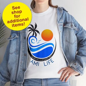 K&ouml;nnte beinhalten: Wei&szlig;es T-Shirt mit einem blauen und orangenen Motiv aus einer Palme, Sonne und Wellen. Der Text "Gulf tides and good vibes AMI LIFE" ist unter dem Motiv aufgedruckt.