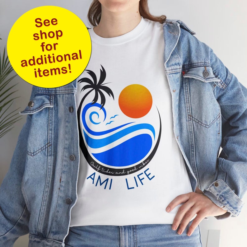 K&ouml;nnte beinhalten: Wei&szlig;es T-Shirt mit einem blauen und orangenen Motiv aus einer Palme, Sonne und Wellen. Der Text "Gulf tides and good vibes AMI LIFE" ist unter dem Motiv aufgedruckt.