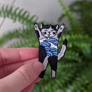 Könnte beinhalten: Gestickter Bügel-Patch mit einem schwarz-weißen Kätzchen mit blauen und weißen Streifen, die Pfoten in die Luft gestreckt.