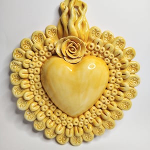 Op de afbeelding: Een gele keramische hartvormige wanddecoratie. Het hart is omgeven door een decoratieve rand van kleine, afgeronde vormen en krullen. Bovenop een roos en vlammen. Het stuk heeft een warme, crèmekleurige gele kleur.