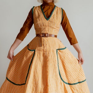 以下が含まれることがあります： 黄色の白のギンガムチェックのドレスに緑色のトリムと茶色のベルトが付いています。ドレスはVネックで、スカートはフルスカートです。