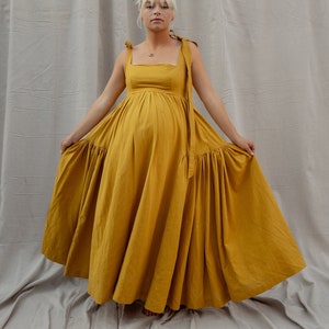 以下が含まれることがあります： スクエアネックと細いストラップのマスタードイエローのロングドレスを着た女性。ドレスはギャザーの入ったスカートで、軽量な生地で作られています。