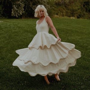 以下が含まれることがあります： クリーム色のティアードドレス、流れるようなスカート付き。 ドレスはフィットしたボディスとスパゲッティストラップが付いています。 モデルは背景に木々のある草地の中に立っています。