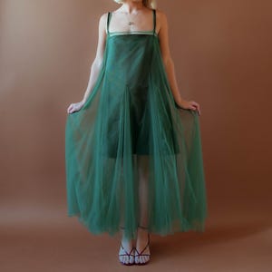 May include: A dark green dress with a sheer overlay. The dress has a square neckline and thin straps. The model is wearing a necklace and sandals. The background is a solid brown colour.