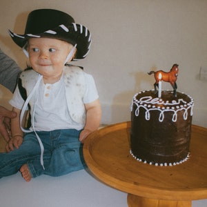 May include: A young child wearing a black and white cowboy hat and vest sits in front of a chocolate cake with white frosting and a small brown horse figurine on top. The cake is on a wooden cake stand.