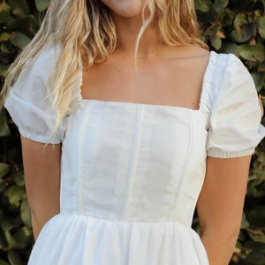 以下が含まれることがあります： スクエアネックとパフスリーブの白いドレス。ドレスはフィットしたボディスと流れるようなスカートを持っています。