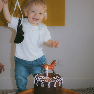 May include: A young child wearing a white shirt and blue jeans stands in front of a chocolate cake with white frosting. The cake is decorated with a small brown horse figurine and a lit candle. The cake says "Oliver" on the frosting.