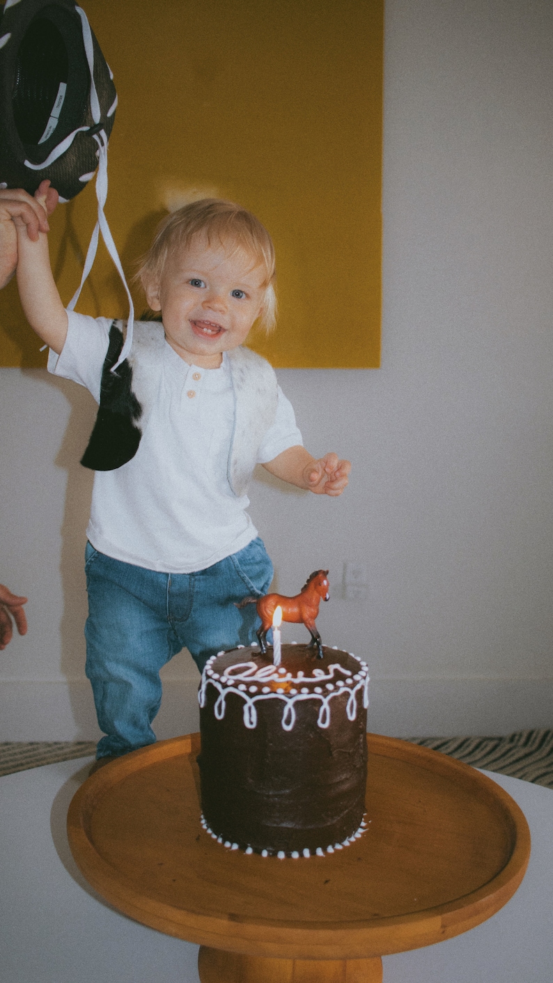 May include: A young child wearing a white shirt and blue jeans stands in front of a chocolate cake with white frosting. The cake is decorated with a small brown horse figurine and a lit candle. The cake says "Oliver" on the frosting.