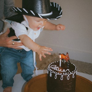 May include: A young child wearing a black and white cowboy hat and a white shirt with a black and white pattern blows out a candle on a chocolate cake decorated with white frosting and the name "Oliver". A small brown horse figurine sits on top of the cake.