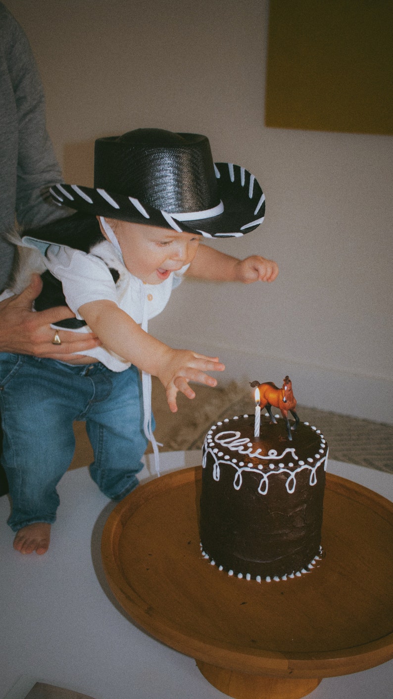May include: A young child wearing a black and white cowboy hat and a white shirt with a black and white pattern blows out a candle on a chocolate cake decorated with white frosting and the name "Oliver". A small brown horse figurine sits on top of the cake.