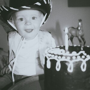 May include: A young child wearing a black cowboy hat with white trim sits in front of a chocolate cake with white frosting and a single lit candle. A small toy horse is on top of the cake.
