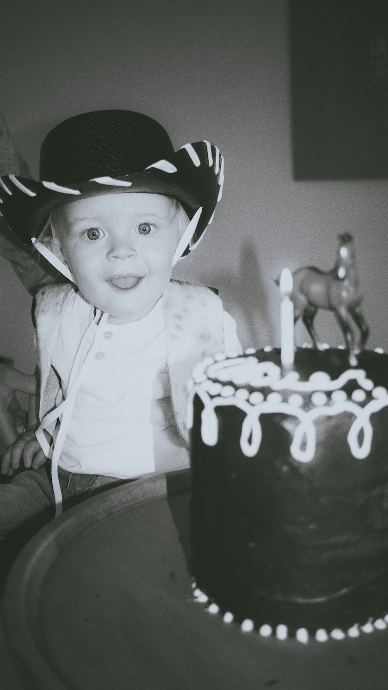 May include: A young child wearing a black cowboy hat with white trim sits in front of a chocolate cake with white frosting and a single lit candle. A small toy horse is on top of the cake.
