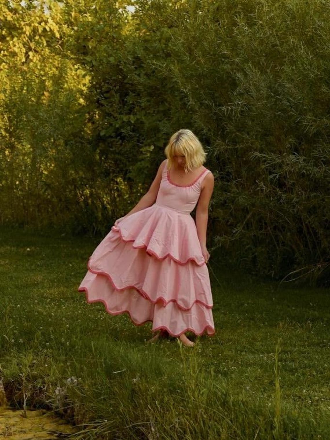 May include: A light pink dress with a tiered skirt and a scoop neckline. The dress has a darker pink trim along the edges of the tiers. The dress is worn outdoors in a grassy area with trees in the background.
