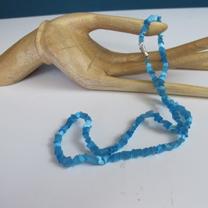 May include: A light blue necklace made of square-shaped beads. The necklace is on a wooden hand.
