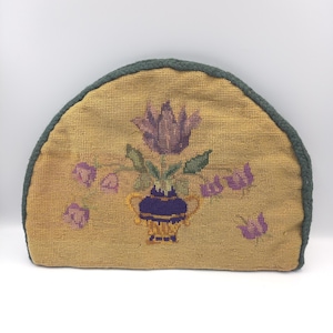 Op de afbeelding: Een halve cirkel theemuts met een bloemenpatroon in kruissteek. De theemuts heeft een gele achtergrond met een paarse tulp en andere paarse bloemen in een blauwe vaas. De randen zijn afgewerkt met een groen touw.