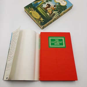 Puede incluir: Un libro de tapa dura rojo titulado "The Saturday Book" con un borde verde alrededor del título. El libro está abierto para revelar páginas en blanco.