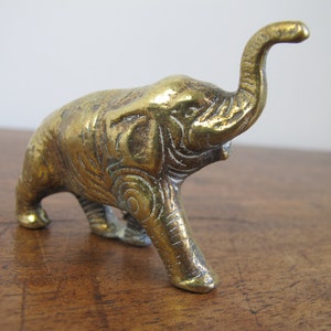 May include: A small, gold-colored brass elephant figurine with its trunk raised in the air. The elephant has intricate details carved into its body.