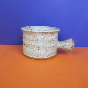 Puede incluir: Una taza pequeña de cerámica beige con un esmalte moteado y un asa marrón. La taza está sobre una superficie naranja.
