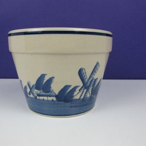 May include: A white ceramic planter with a blue and white windmill design. The planter has a blue rim and a blue base.