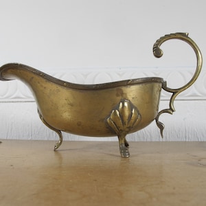 May include: A vintage brass gravy boat with a decorative handle and three legs. The boat has a scalloped edge and a floral design on the side.