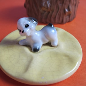 Może przedstawiać: Ceramiczna pokrywka z żółtą glazurą i małą biało-czarną figurką psa na górze. Pokrywka ma efekt spękań glazury.