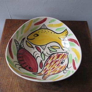 Puede incluir: Un cuenco de cerámica blanca con forma triangular presenta un diseño estilizado de tres peces. Los peces están pintados en amarillo, rojo y verde con contornos negros. El cuenco está decorado con un fondo de hojas amarillas, rojas y verdes.