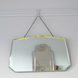 Pode incluir: Um espelho vintage biselado com uma borda amarela pendurado em uma corrente. O espelho tem uma forma geométrica e reflete um teto branco e uma luminária.