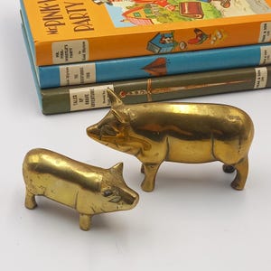 Könnte beinhalten: Zwei Messing-Schwein-Figuren, eine große und eine kleine, sind vor einem Stapel alter Kinderbücher zu sehen. Die Bücher haben farbenfrohe Einbände mit Titeln wie "Mr. Pink-Whistle's Party" und "Tales of Brave Adventure."