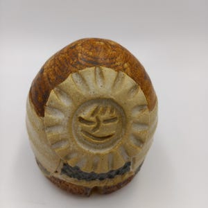 May include: A rounded ceramic object with a textured brown and cream finish. The front features a raised, circular sun-like emblem with carved facial features. The base has a dark, textured band, and the overall surface shows visible clay markings.